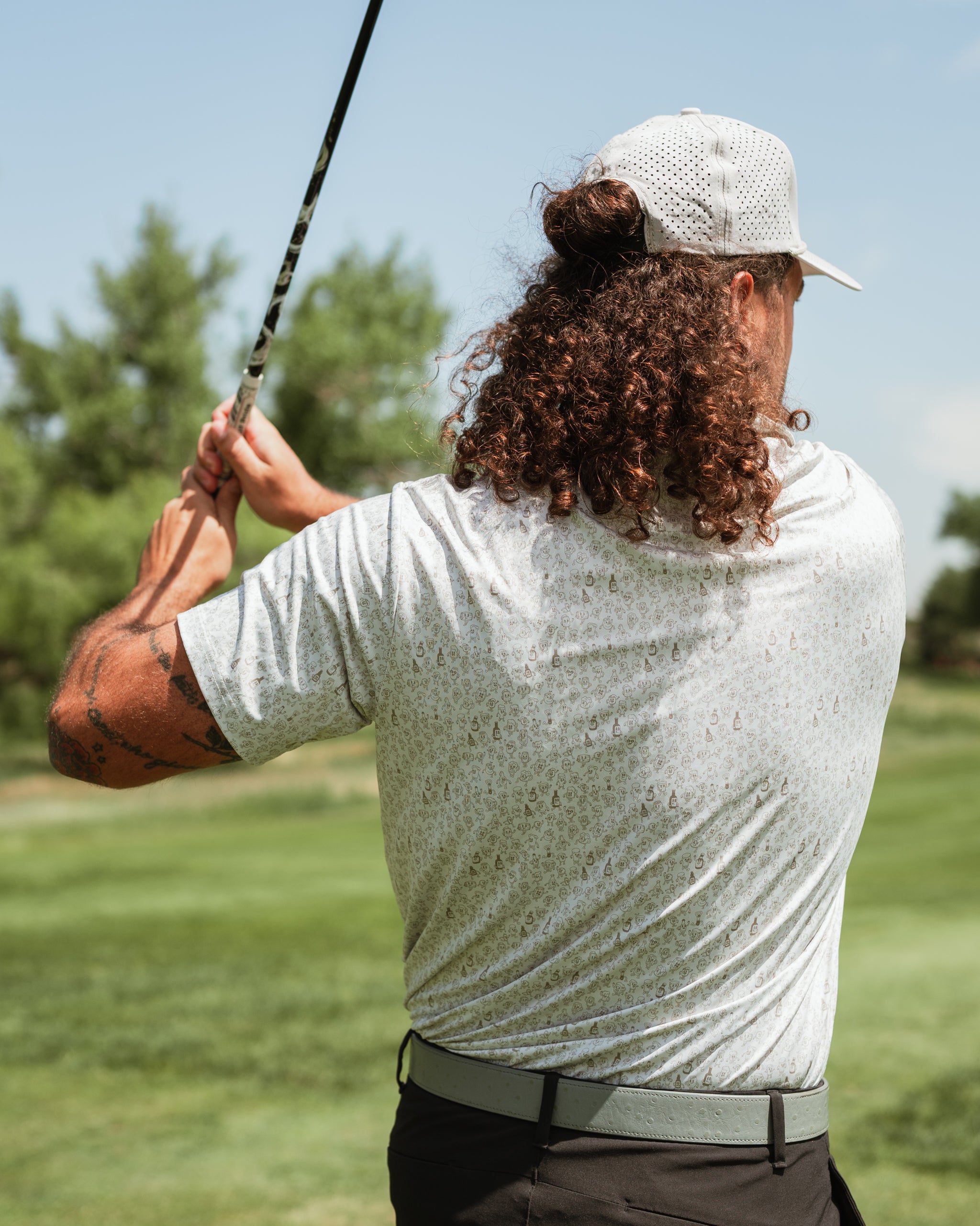 A golfer with curly hair swings a club, wearing a light gray patterned shirt on a sunny course.