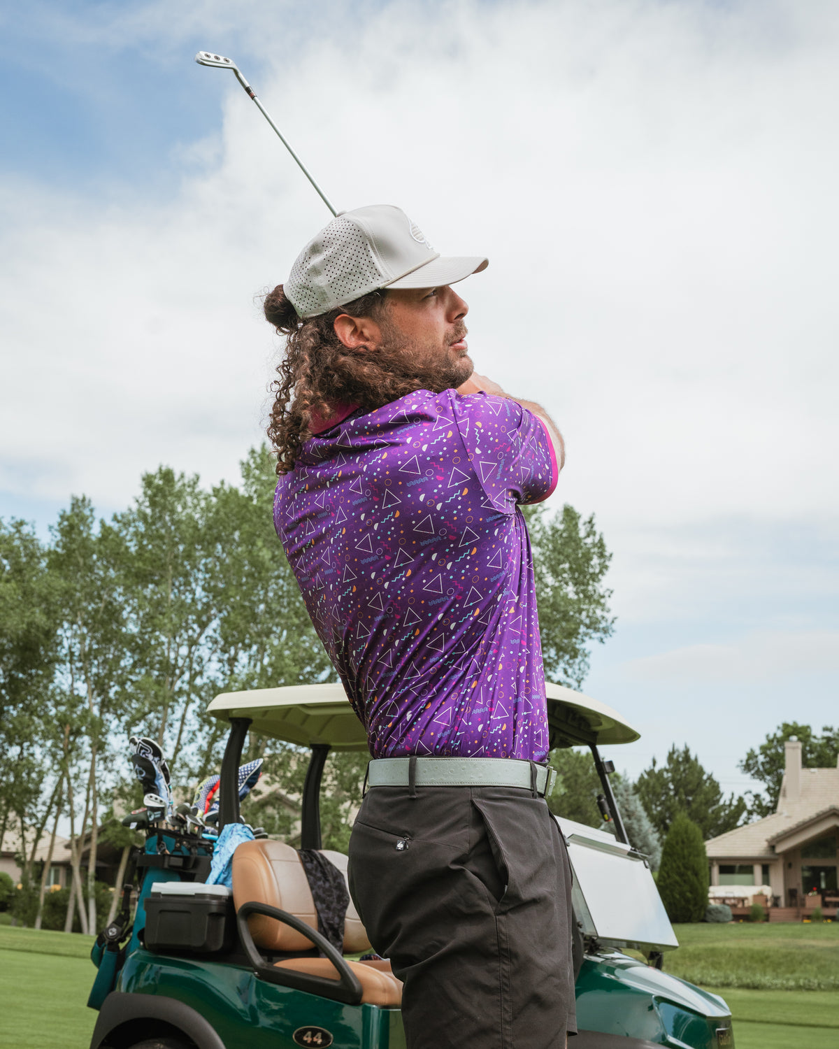 A golfer in a vibrant purple Eighties Arcade polo shirt swings a club on the course, with a golf cart in the background.