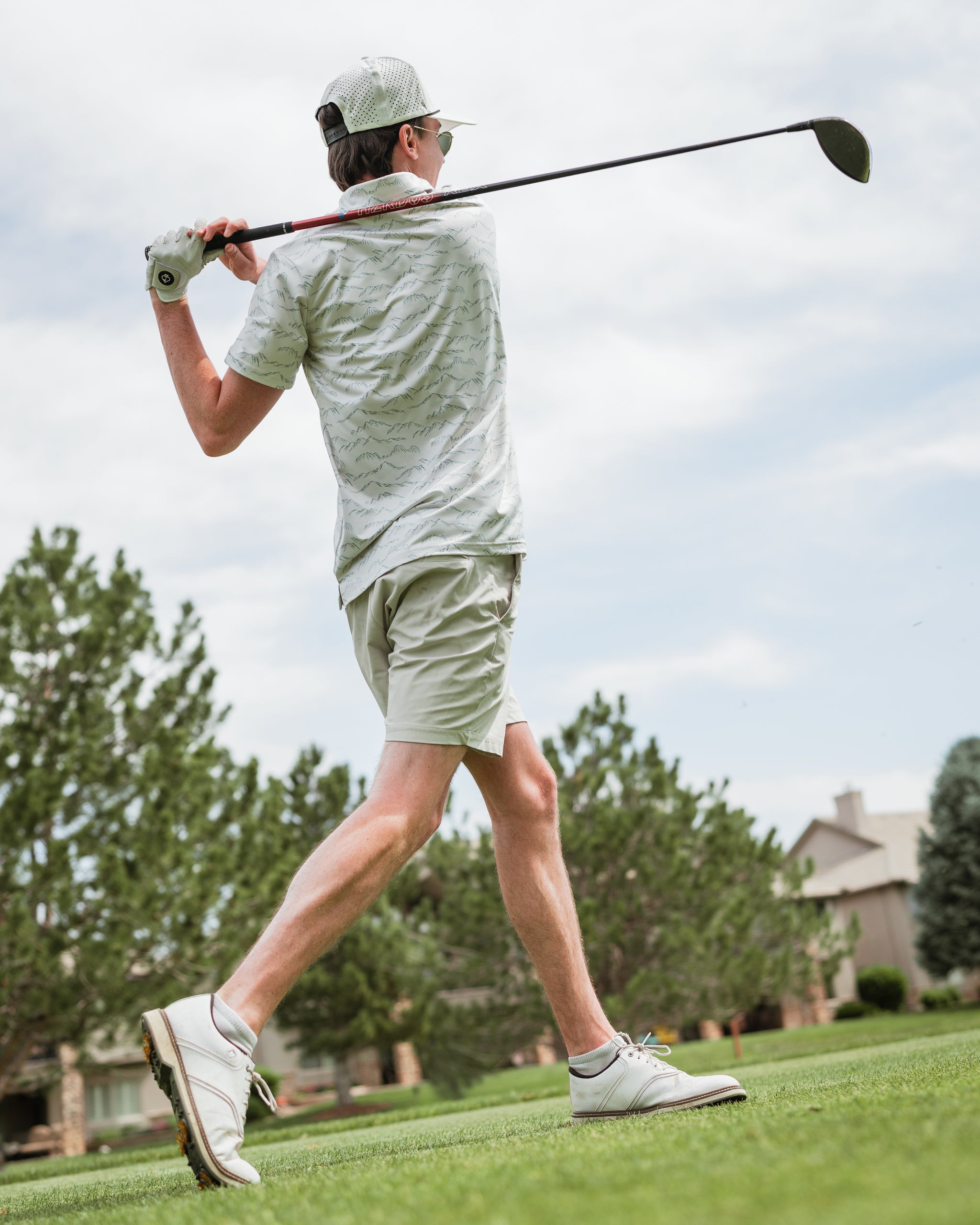 A golfer swings a driver on the course, wearing a light green patterned shirt and beige shorts, showcasing a stylish golf look.