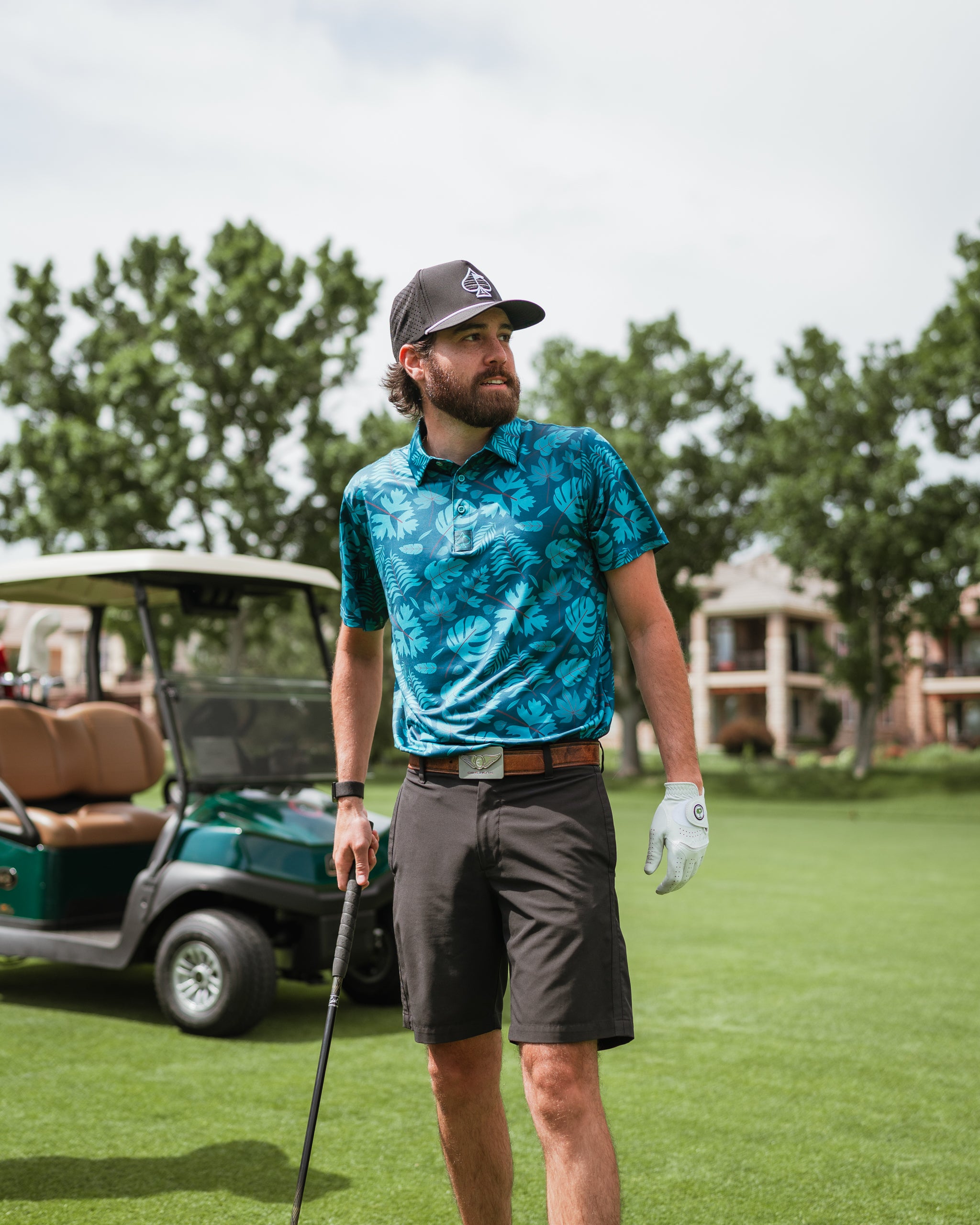 Man wearing a vibrant blue floral golf polo and shorts, standing on a golf course near a golf cart.