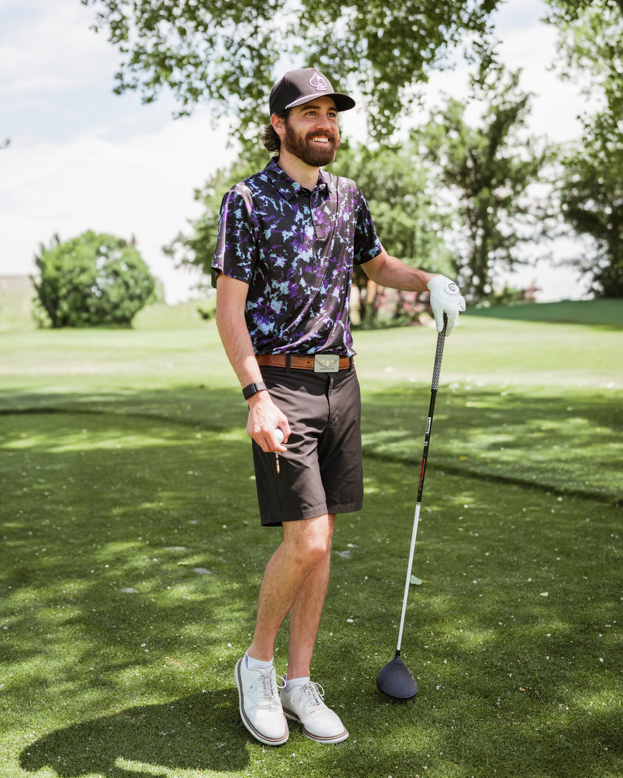 A golfer wearing a floral-patterned polo shirt and black shorts stands on the course, holding a driver.