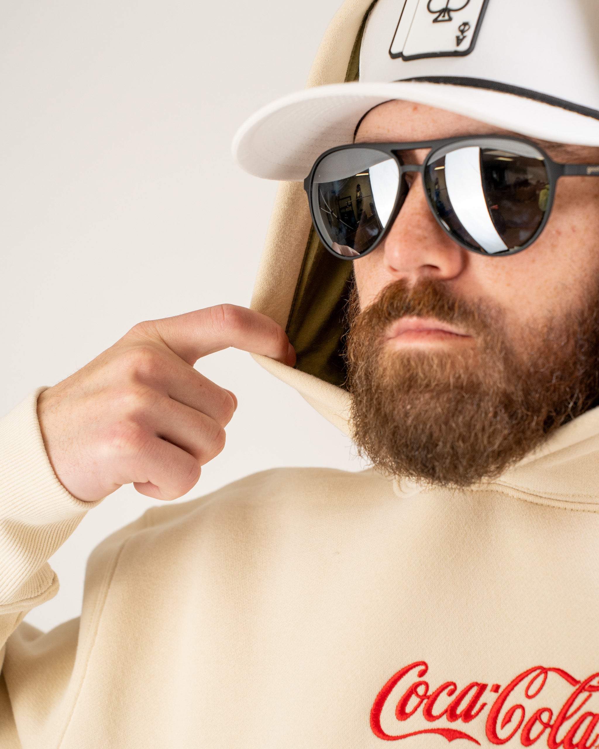 A person wearing a beige Coca-Cola hoodie, adjusting the hood while sporting sunglasses and a white cap.