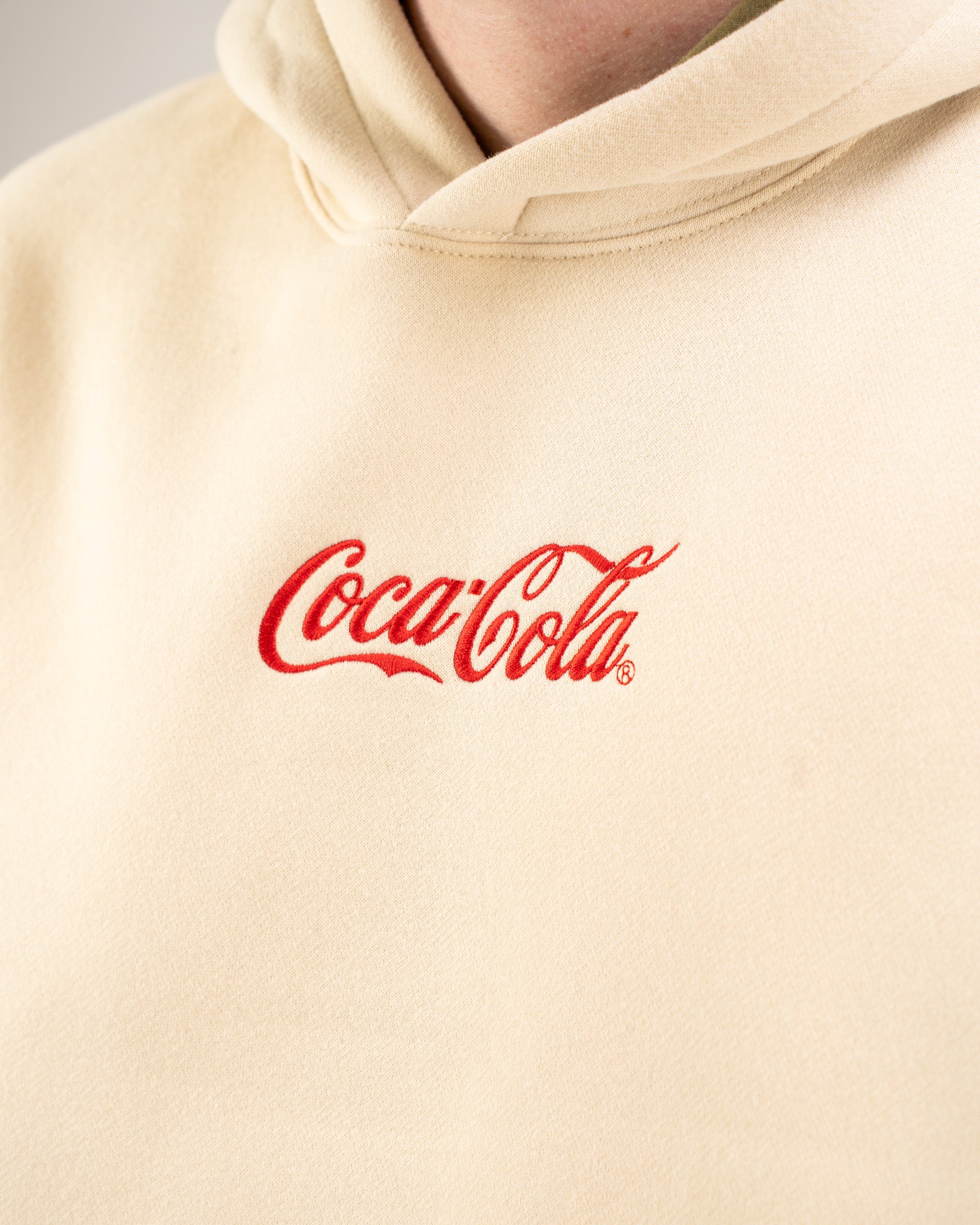 Beige lifestyle hoodie featuring the classic Coca-Cola logo embroidered in red on the chest. Perfect for casual wear.
