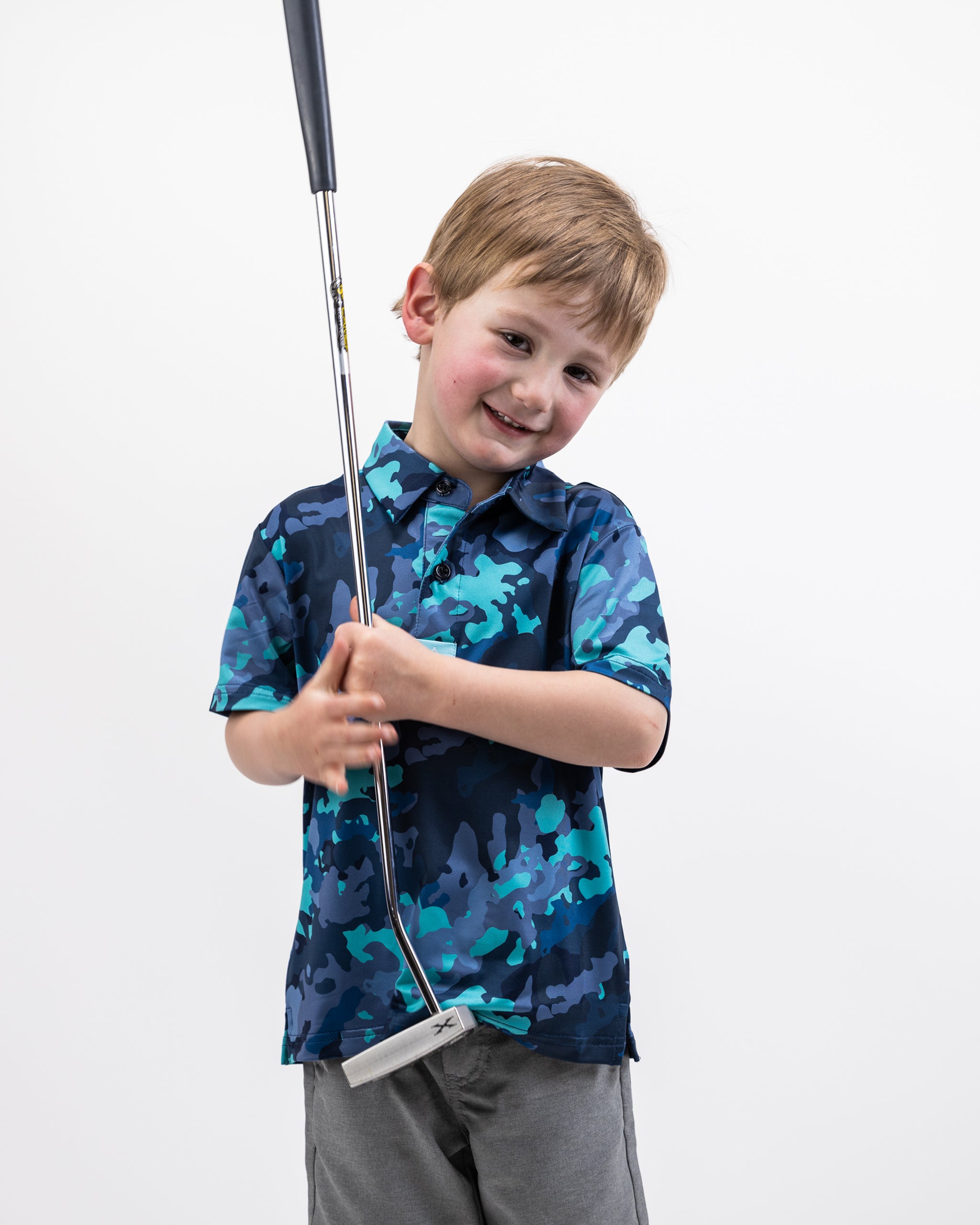 Young boy smiling in a blue electric camo golf shirt, holding a golf club against a plain white background.
