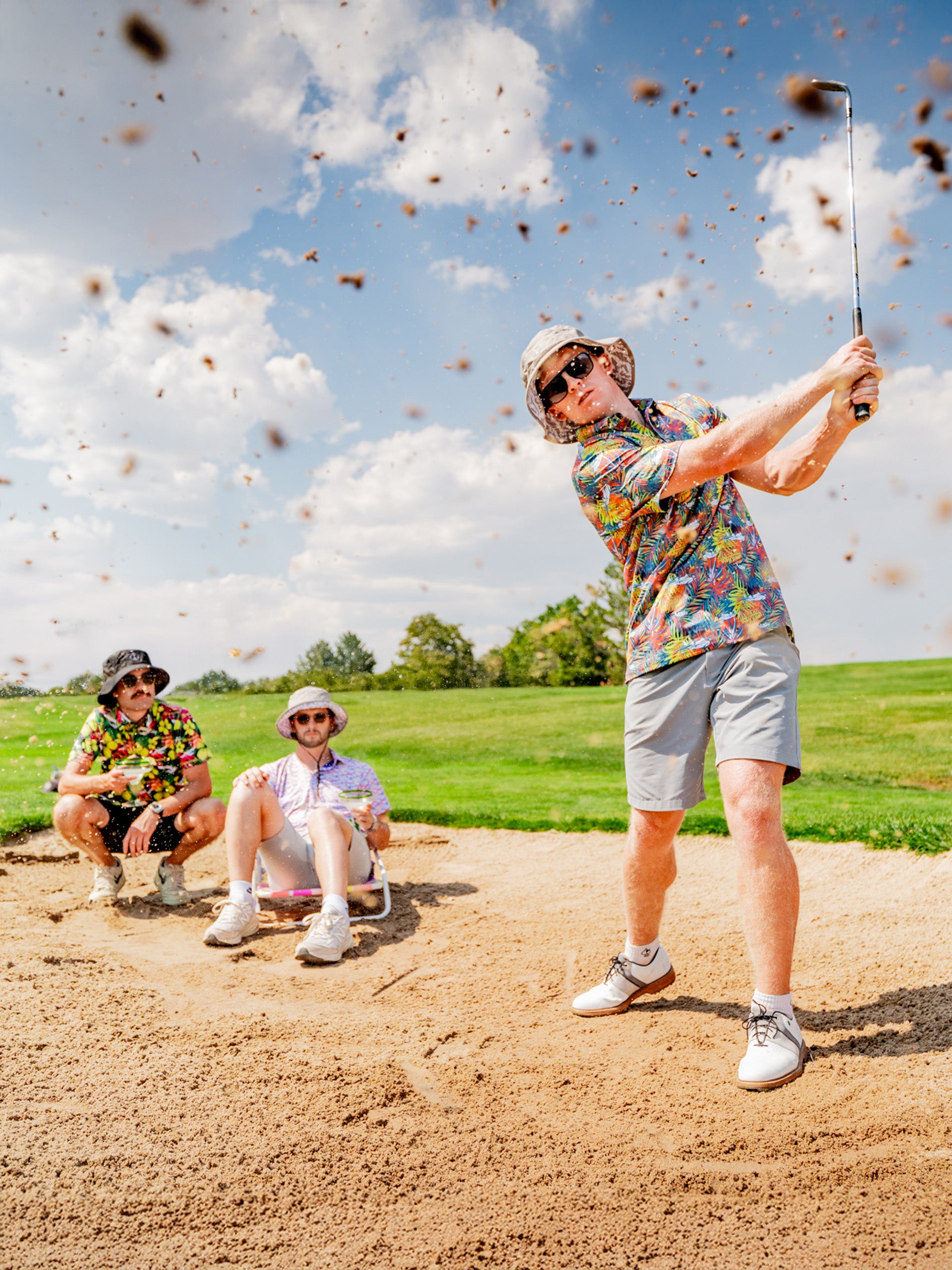 A golfer swings a club in a sandy area while two friends relax nearby, all wearing vibrant tropical shirts.