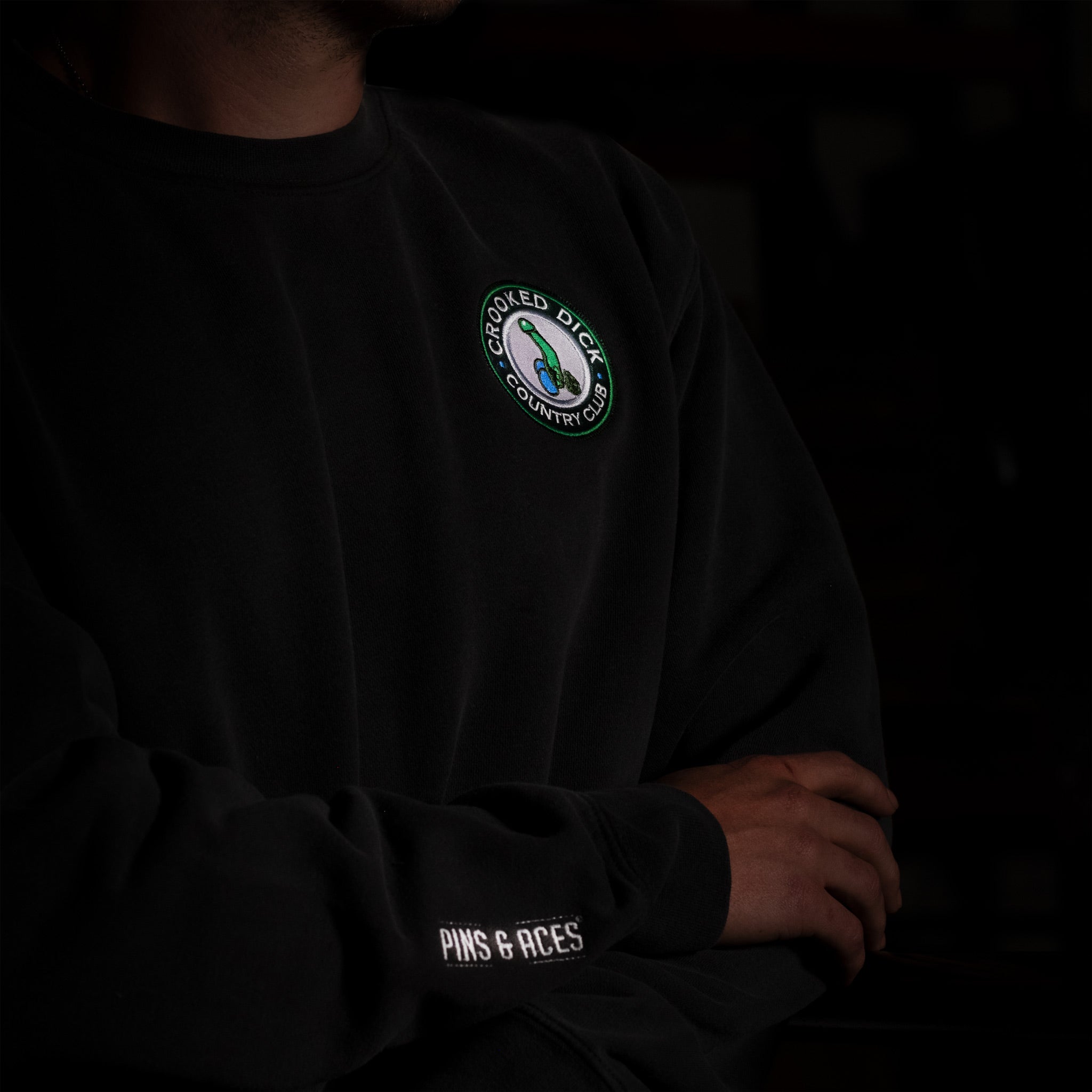 Black sweatshirt with "Crooked Dick Country Club" embroidered patch and "Pins & Aces" on sleeve, for bold golf style.