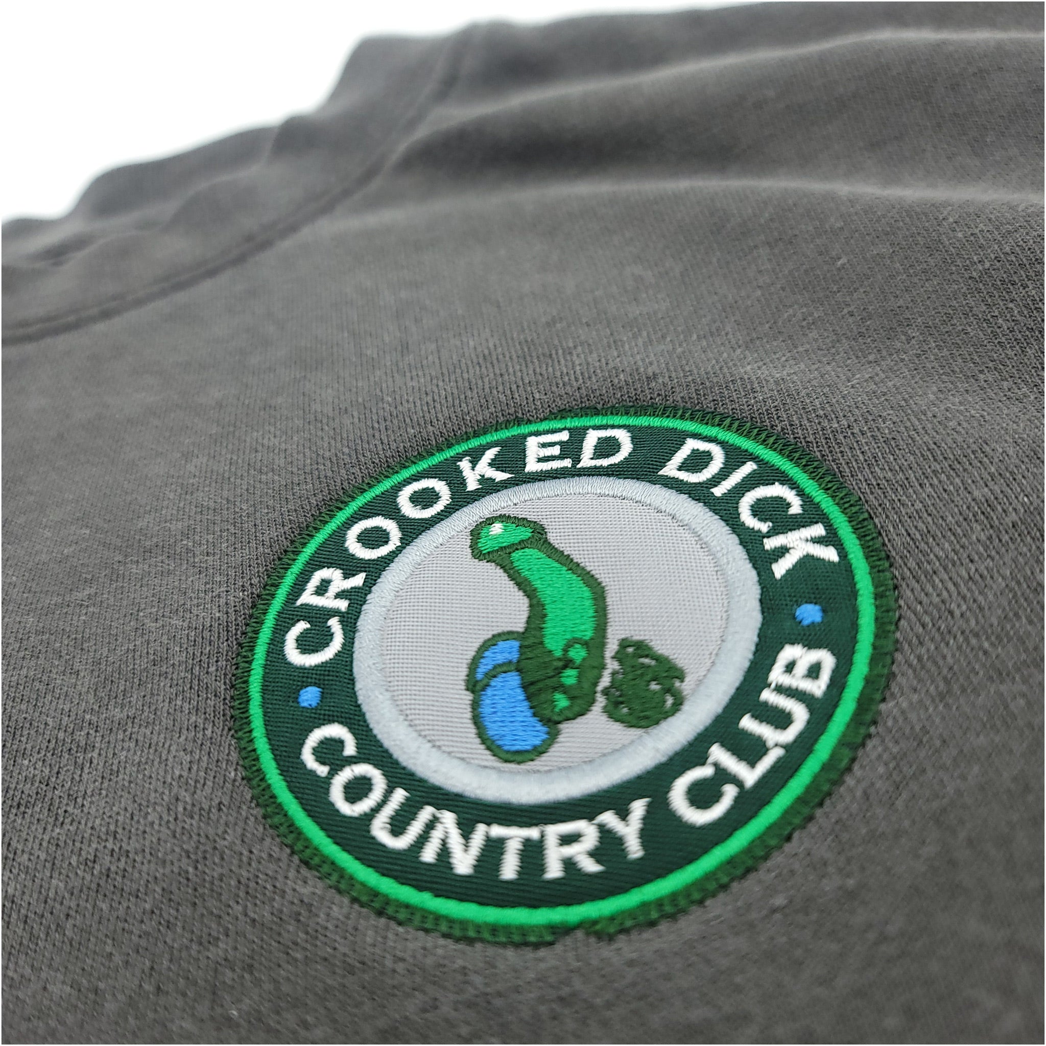 Close-up of a gray golf shirt with a colorful embroidered "Crooked Dick Country Club" logo featuring a green golf club and...